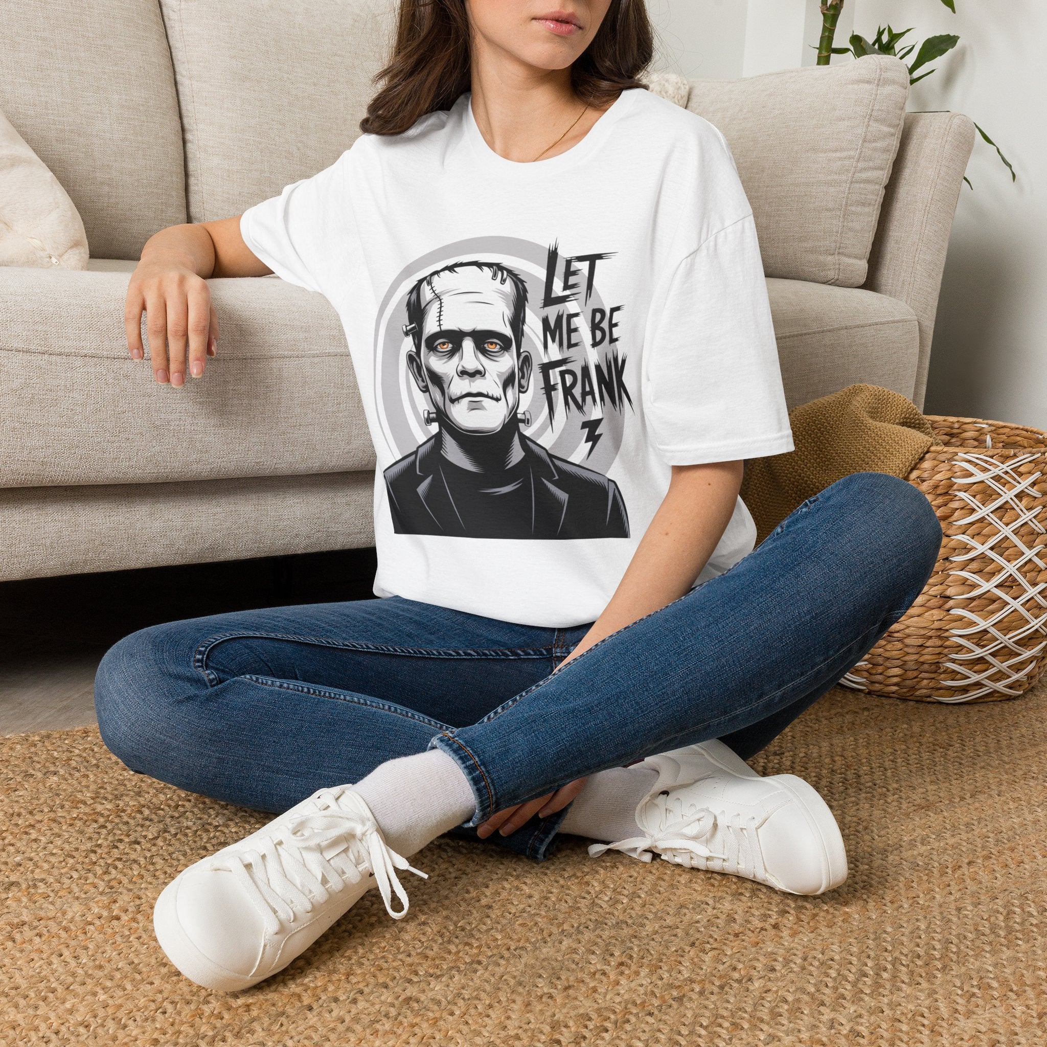 A woman sitting cross-legged on a woven rug in front of a beige sofa, wearing a white graphic T-shirt with a black-and-gray Frankenstein portrait and the text 'LET ME BE FRANK'.