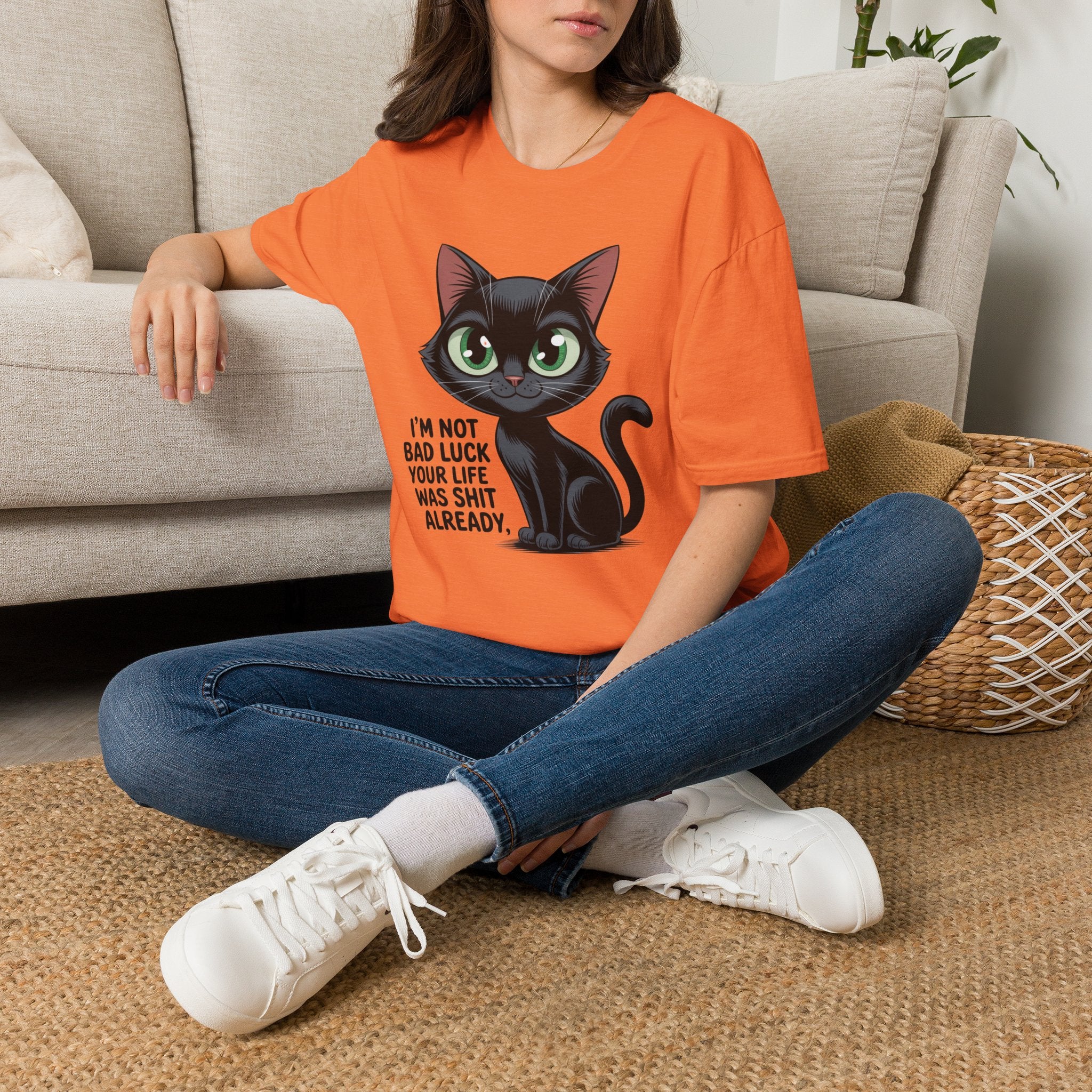 A woman sitting cross-legged on a woven rug in front of a beige sofa, wearing an orange graphic T-shirt with a black cat illustration and the text 'I'M NOT BAD LUCK YOUR LIFE WAS SHIT ALREADY.' She is also wearing blue jeans and white sneakers.