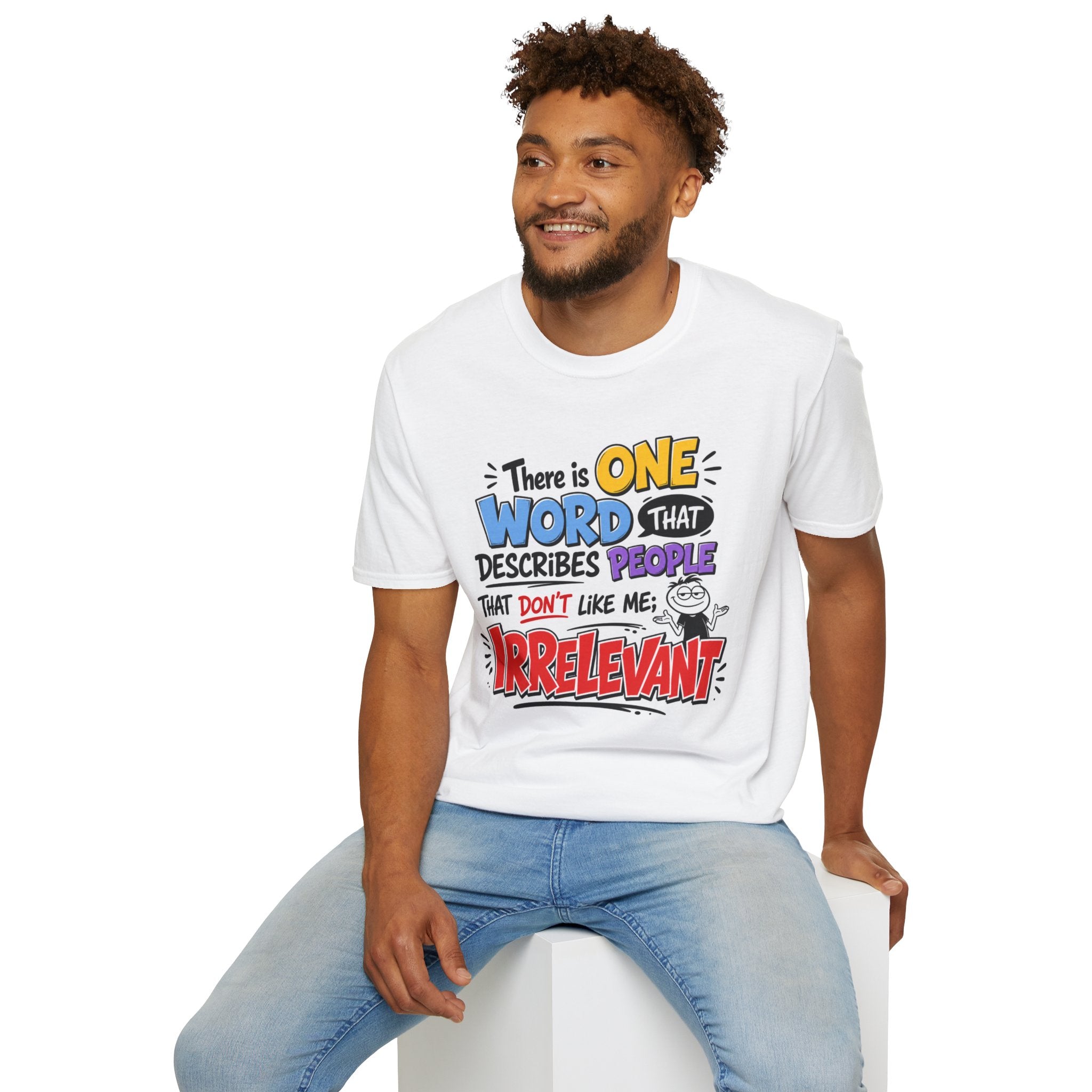 A man wearing a white graphic t-shirt with a colorful text design that reads 'There is ONE WORD that DESCRIBES PEOPLE that don't like me: IRRELEVANT', paired with light blue jeans, sitting on a white block against a white background.