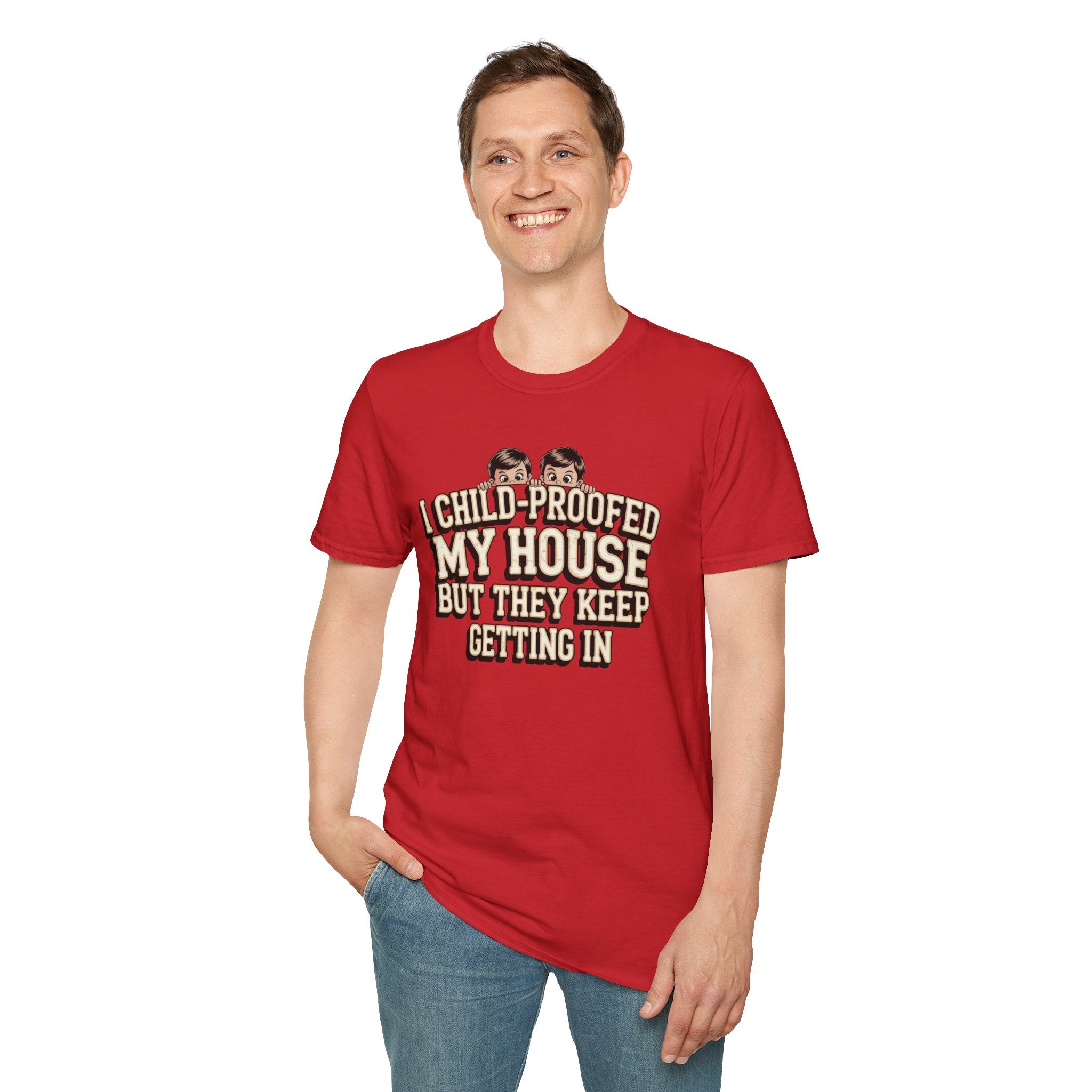 A man wearing a red graphic t-shirt with the text 'I CHILD-PROOFED MY HOUSE BUT THEY KEEP GETTING IN' in beige and black, paired with blue jeans, against a plain white background.