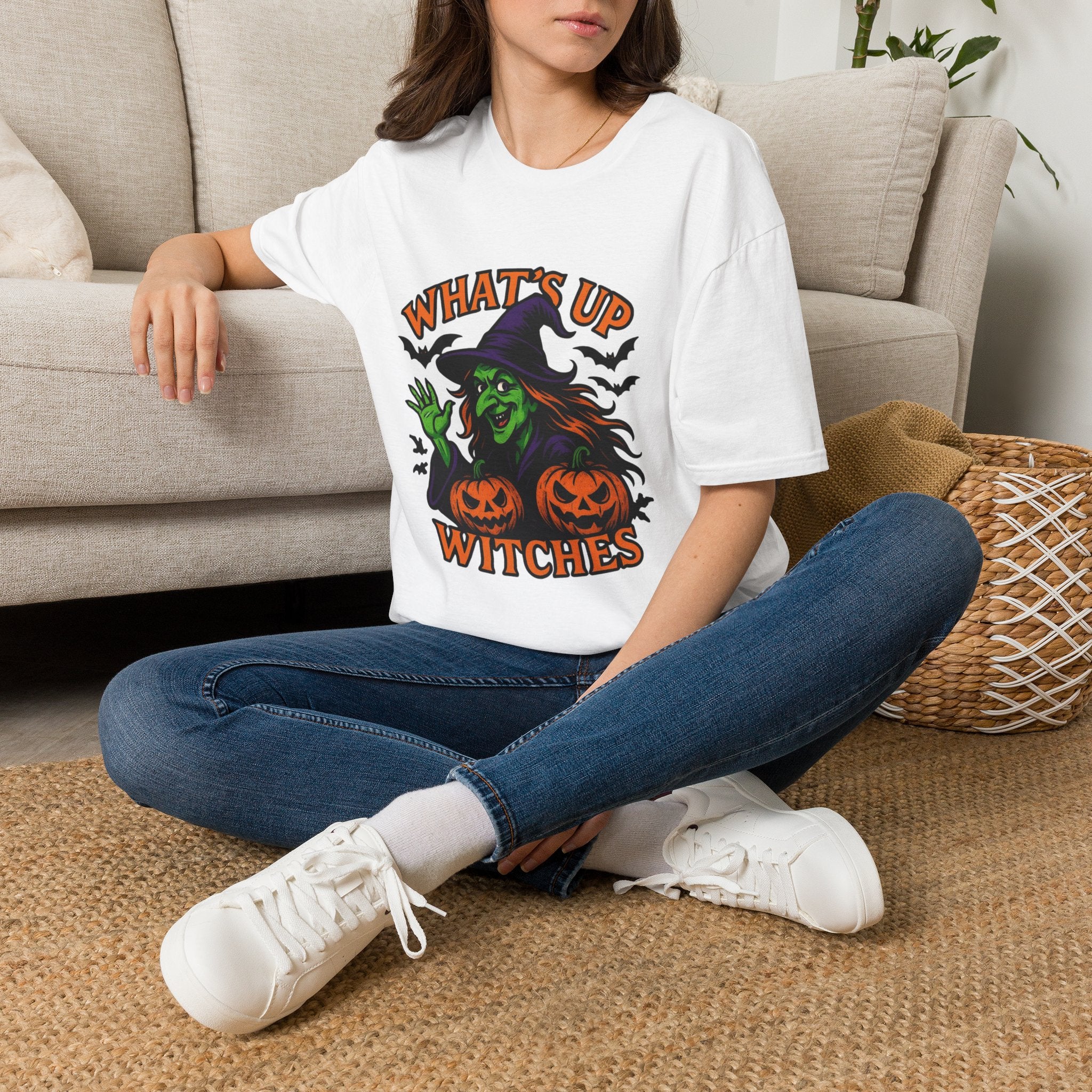 A woman sitting cross-legged on a woven rug in front of a beige couch, wearing a white graphic T-shirt with a Halloween witch design and the text 'WHAT'S UP WITCHES'.