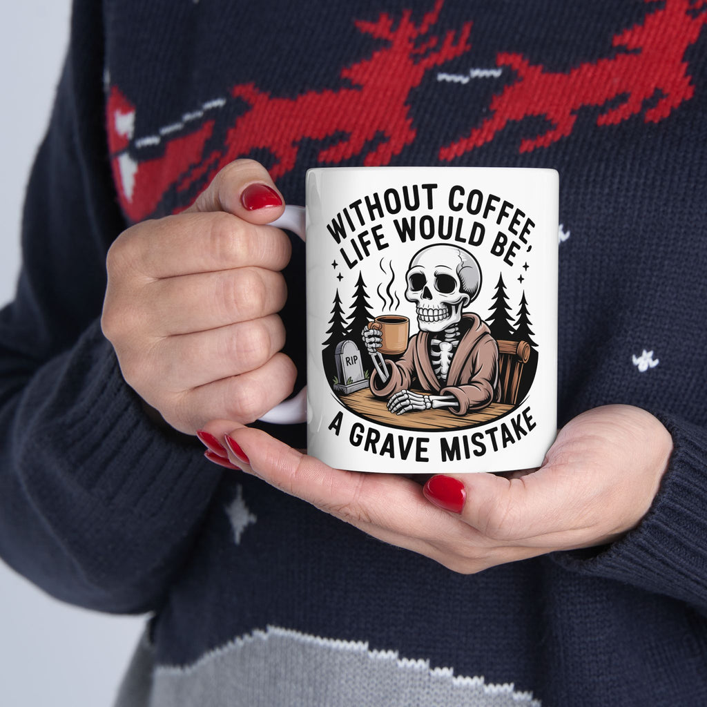 A person holding a white ceramic mug with a humorous graphic of a skeleton in a suit sitting in a grave with a coffee mug, surrounded by the text 'WITHOUT COFFEE LIFE WOULD BE A GRAVE MISTAKE'. The person is wearing a dark navy knit sweater with red and gray patterns.