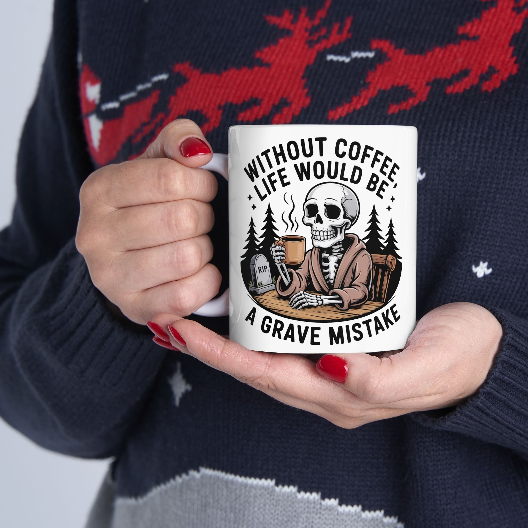 A person holding a white ceramic mug with a humorous graphic of a skeleton in a suit sitting in a grave with a coffee mug, surrounded by the text 'WITHOUT COFFEE LIFE WOULD BE A GRAVE MISTAKE'. The person is wearing a dark navy knit sweater with red and gray patterns.