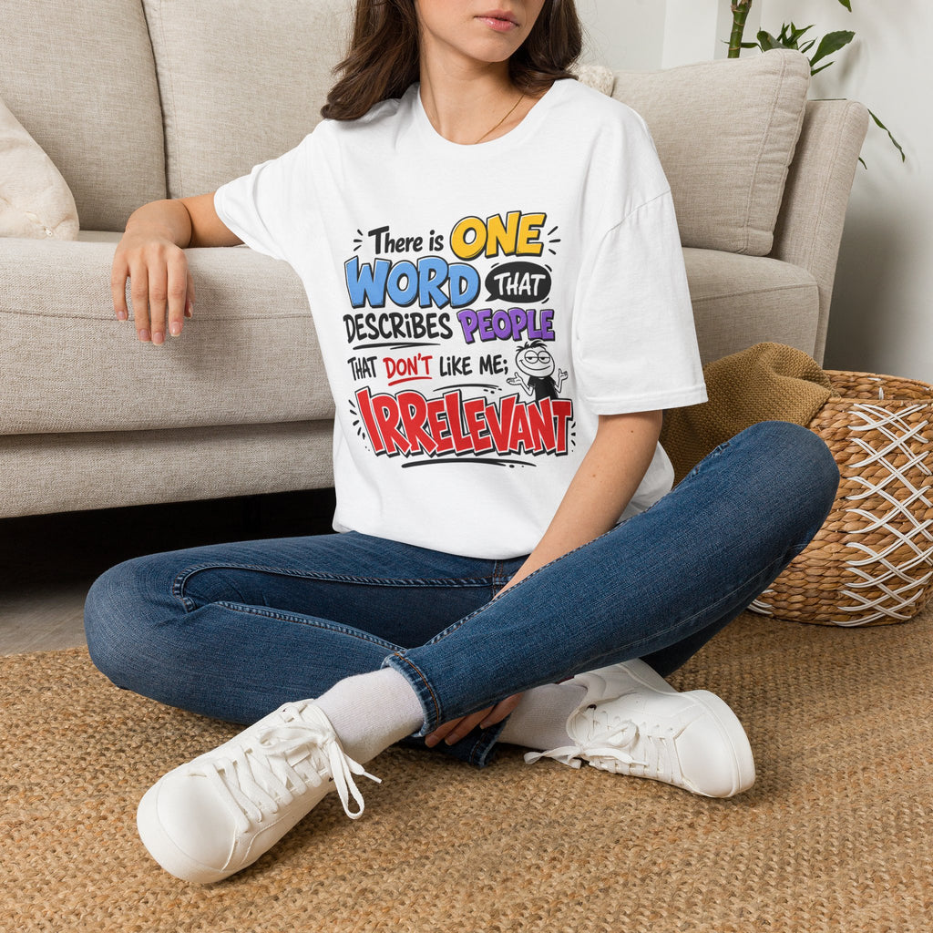 A woman sitting cross-legged on a woven rug in front of a beige sofa, wearing a white graphic T-shirt with colorful text that reads, 'There is ONE WORD that DESCRIBES PEOPLE that DON'T like me: Irrelevant.' She is also wearing blue jeans and white sneakers.