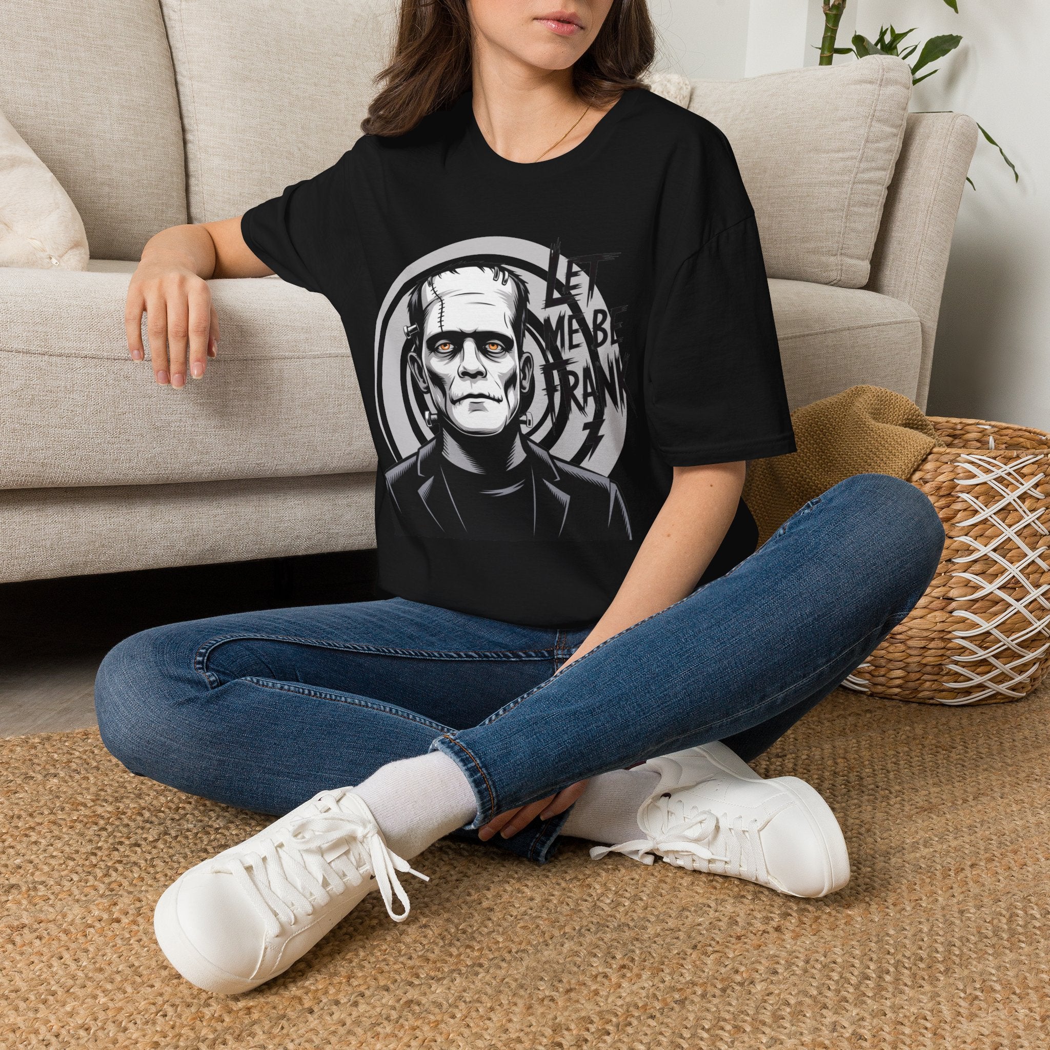 A woman sitting cross-legged on a woven rug in front of a beige sofa, wearing an oversized black graphic T-shirt with a white Frankenstein-style monster face print, blue jeans, and white sneakers.